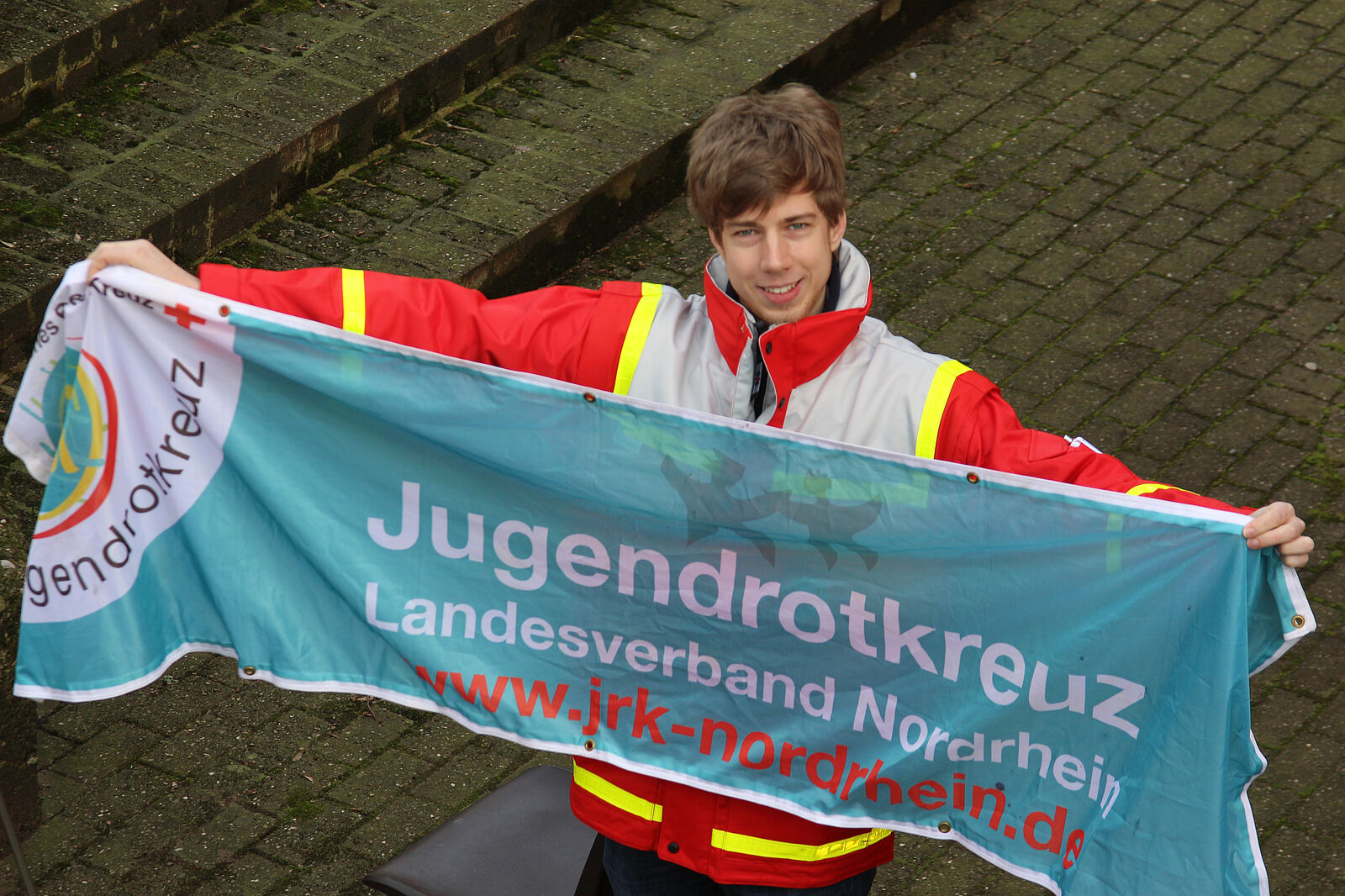 Bild: Ein junger Mensch hält einen Banner. Auf dem Banner ist der Schriftzug "Jugendrotkreuz Landesverband Nordrhein" und die Website www.jrk-nordrhein.de zu lesen.